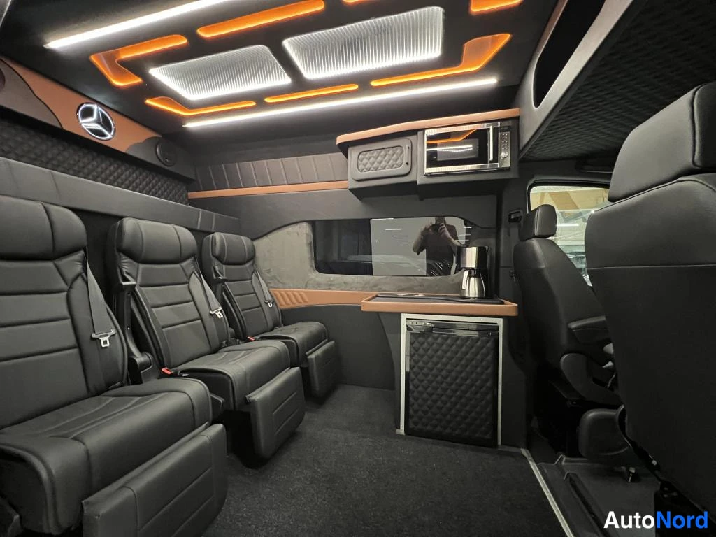 Muu: Kuva kuormatilan takaa Mercedes-Benz Sprinter -autossa: neljä mustaa istuinta, mikroaaltouuni, jääkaappi ja kahvinkeitin