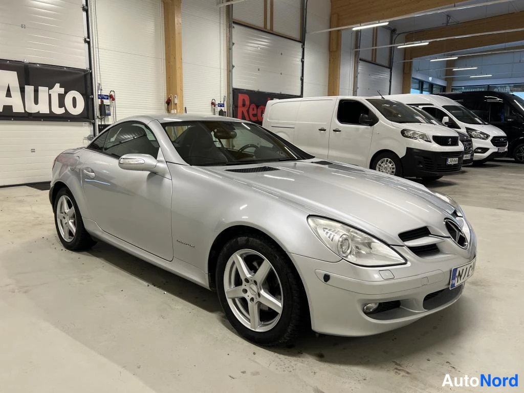 Etuviisto, nokka oikealle: Auton keula oikealta kolmessa neljänneksessä: hopea Mercedes-Benz SLK hallissa.
