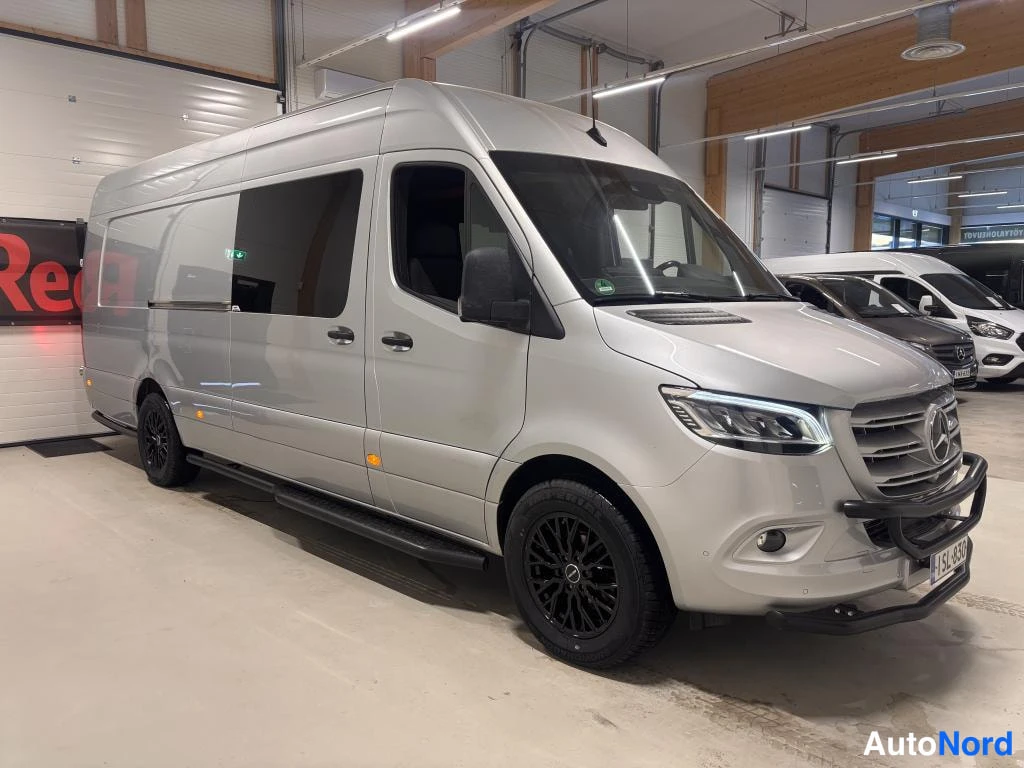 Etuviisto, nokka oikealle: Hopeinen Mercedes-Benz Sprinter -pakettiauto kolmivartsaasta edestä oikealta kuvattuna hallissa, mustat vanteet ja puskurikaari.