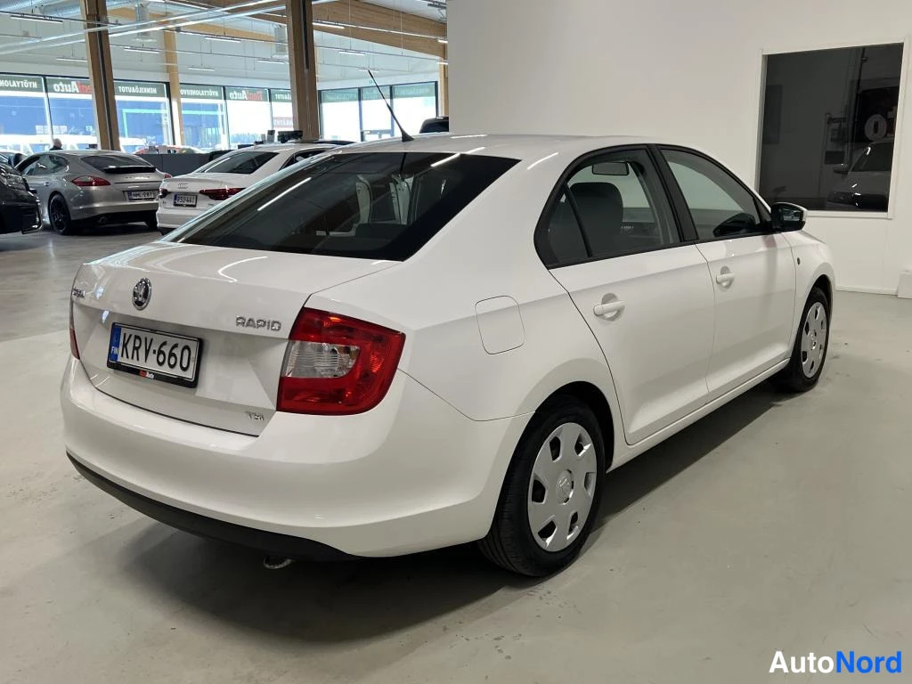Takaviisto: Valkoisen Skoda Rapid -sedan takakulmanäkyvää sisätiloissa, rekisteritunnus, hopeavanteet ja sivupeilit näkyvät.