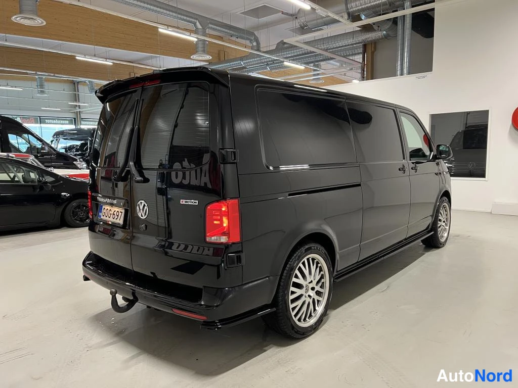Takaviisto: Mustan pakettiauton takakulma kuvattuna sisätilassa; rekisterikilpi, hakenut vetokoukku takapuskurissa.