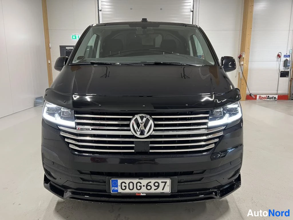 Edestä: Mustan pakettiauton etunäkymä suoraan edestä; kromilistainen etusäleikkö, VW-merkki, 4MOTION-merkki ja rekisterikilpi.