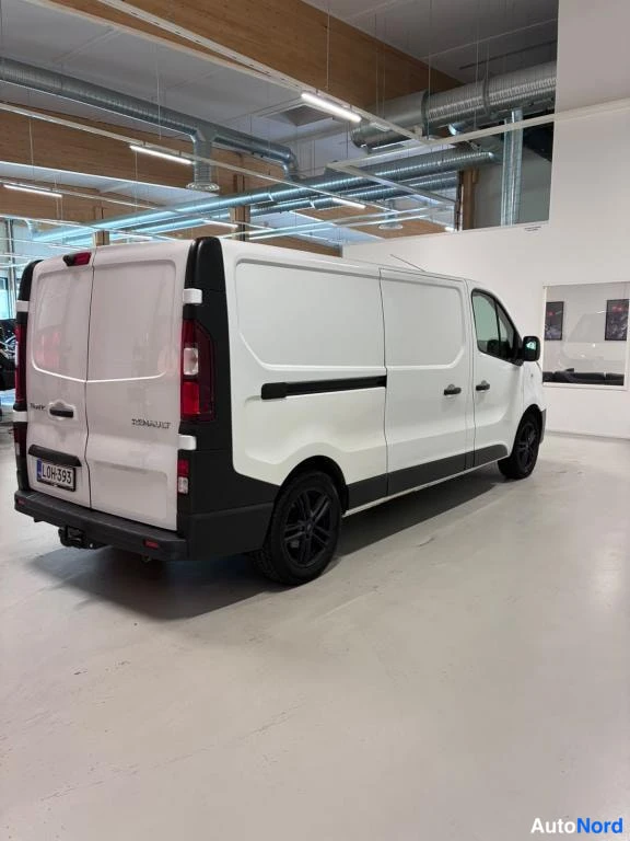 Takaviisto: Valkoinen Renault Trafic -pakettiauto takaa viistosti sisätiloissa: takapari-ovet auki-asennossa näkyvät takavaloineen, musta alareuna ja mustat pyörät.
