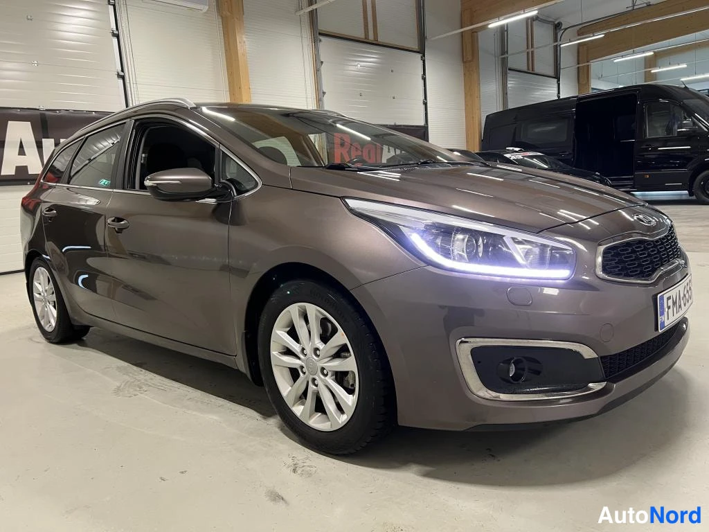 Etuviisto, nokka oikealle: Kia-farmariauto kolmisivukulmassa etuosalla oikealle; ruskeanharmaa kori, hopeiset vanteet ja palaneet etvalot autotallissa.