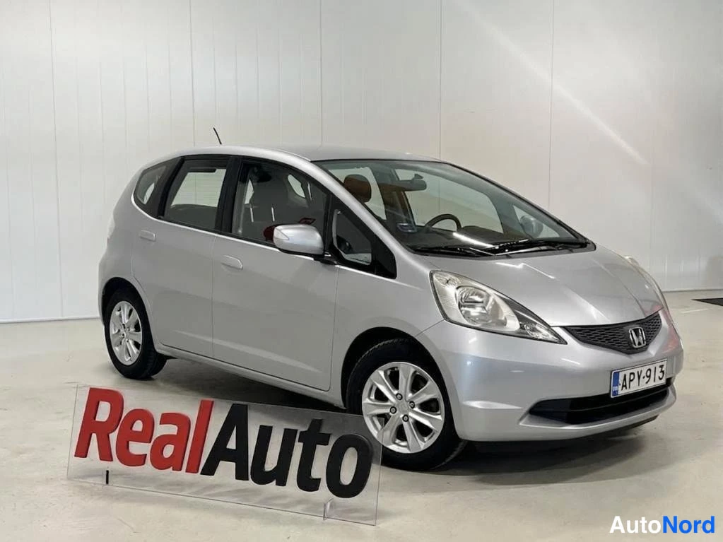 Etuviisto, nokka oikealle: Hopeanvärinen Honda Jazz etuviistosta oikealta, rekisterikilpi, sisätiloissa valkoista seinää vasten.
