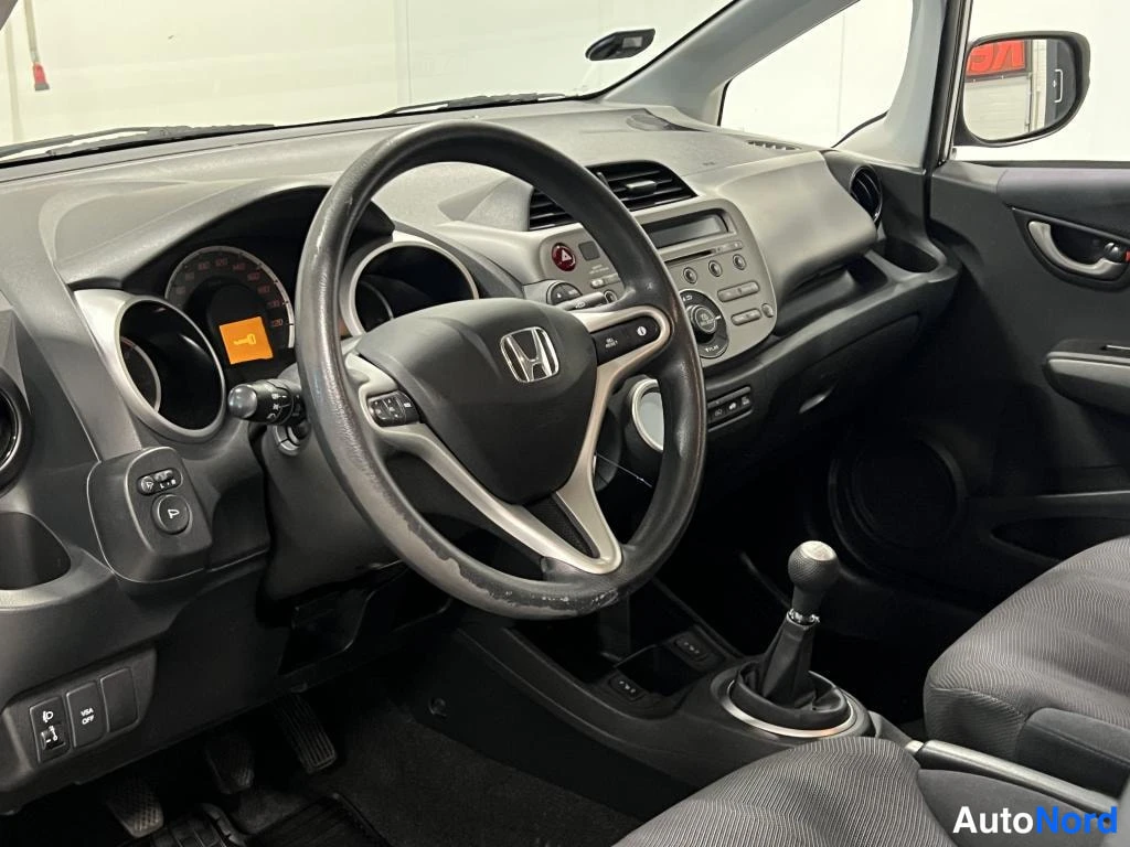 Muu: Honda Jazzin kojelauta ja ohjauspyörä kuljettajan näkökulmasta, manuaalivaihteen vaihtekahva keskellä.