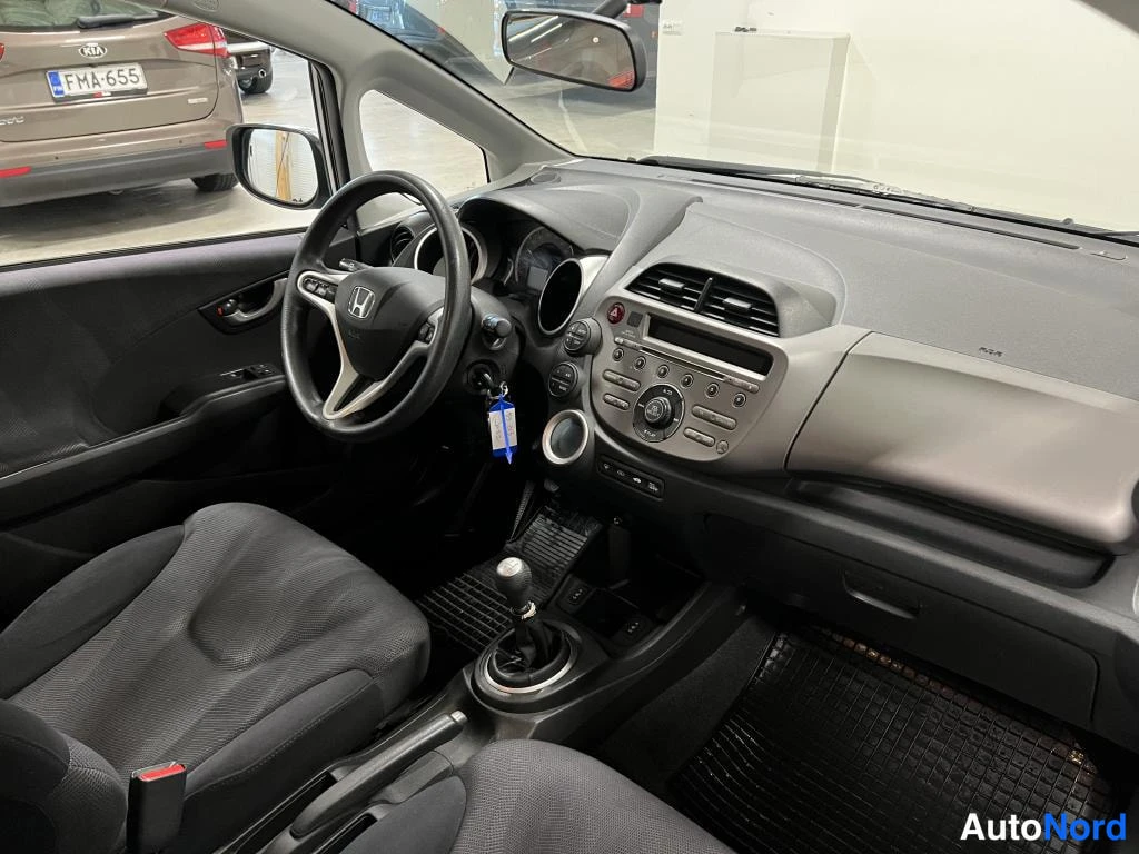 Muu: Honda Jazzin matkustamo kuljettajan puolelta: kojelauta, ohjauspyörä, manuaalivaihteen vaihtekahva ja tummat istuimet.