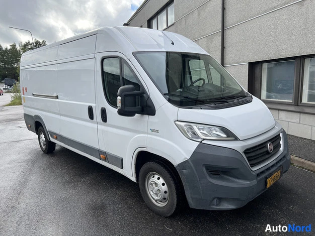 Etuviisto, nokka oikealle: Valkoinen Fiat Ducato -pakettiauto vinosti edestä oikealta kuvattuna rakennuksen vieressä.