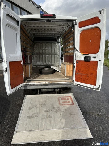 Takaa: Valkoisen pakettiauton takaluukut auki; kuormatilaa verhoaa vaneri- ja oranssi kansiointi, ja lavanosturi on laskettuna asfaltille.