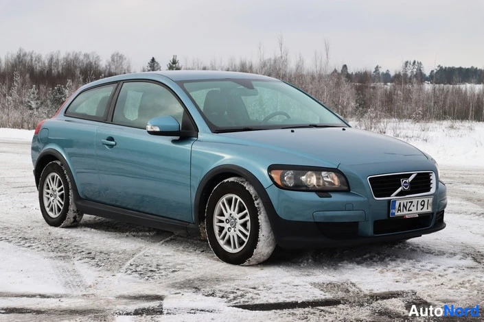 Etuviisto, nokka oikealle: Sinivihreä kaksiovinen henkilöauto etuviistosta lumipeltoa vasten, rekisterikilpi.