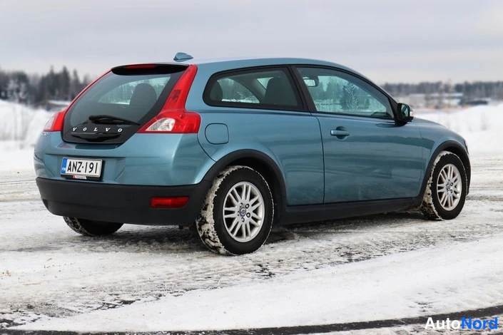 Takaviisto: Auton takakolmasosanäkymä lumisella pihalla; vaaleansininen kolmiovinen automobiili, rekisterikilpi, takaluukussa VOLVO-teksti.