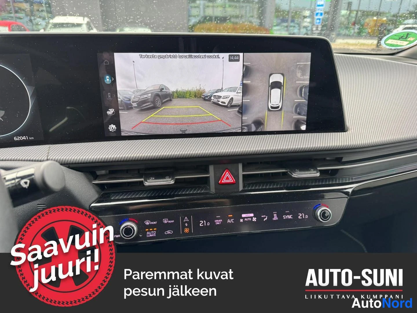 Muu: Auton sisätiloissa kuvattu kojelauta: infotainment-näytöllä ympäristönäkymäkameran ylhäältäpäin kuvattu parkkikuva ja peruutuskameran kuva pysäköintiviivoineen,