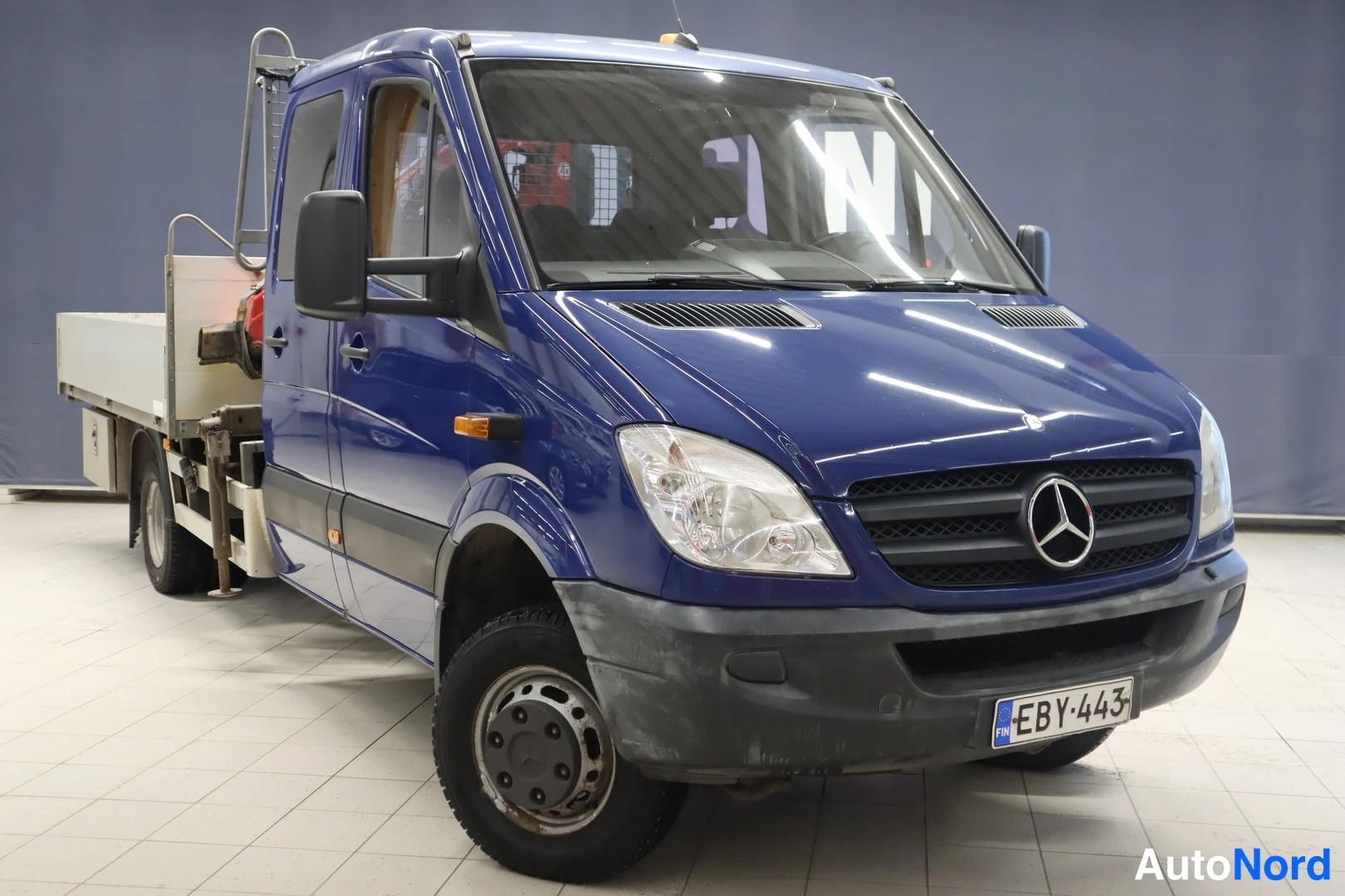 Etuviisto, nokka oikealle: Sininen Mercedes-Benz Sprinter -lavetti-auto kolmesta neljäsosasta etuosalta kuvattuna; rekisterikilpi ja lavalla varusteita.