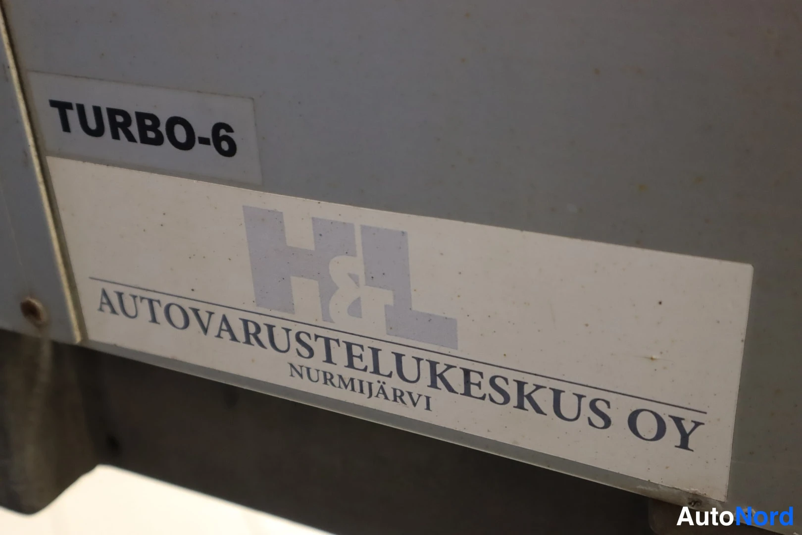 Muu: Valko- ja harmaasävyinen tarra, jossa lukee mustalla TURBO-6 sekä sinisellä H&L:n logo ja teksti AUTOVARUSTELUKESKUS OY, NURMIJÄRVI.