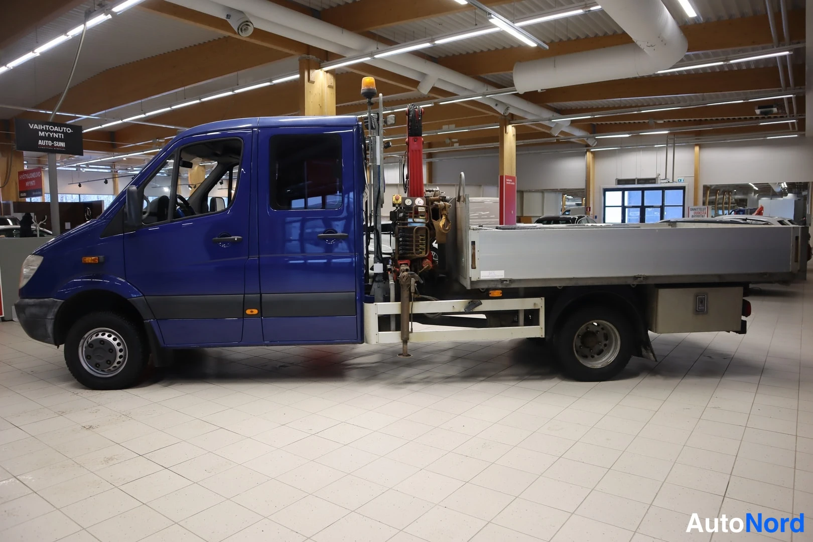Sivulta: Sivukuvassa sininen Mercedes-Benz Sprinter -lava-auto kuormaustilassa; lavalla puristinlaitteisto ja keltainen varoitusvalo.