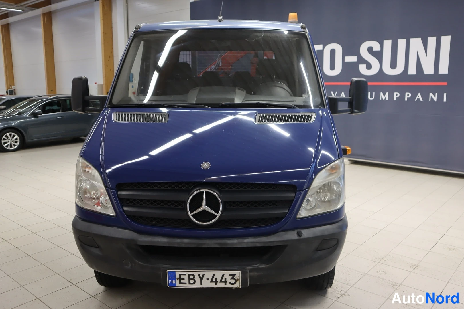 Edestä: Edestä kuvattu sininen Mercedes-Benz Sprinter -lava-auto, rekisterikilpi, myyntihallissa.