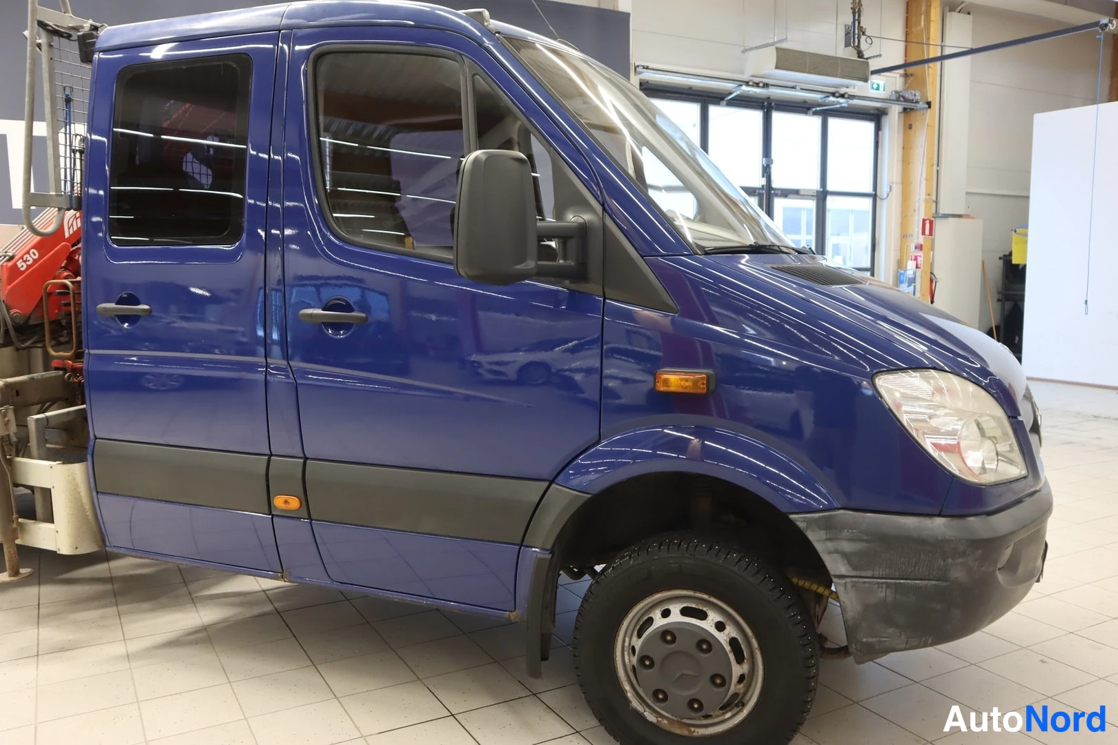 Etuviisto, nokka oikealle: Sininen Mercedes-Sprinter-pakettiauto etukulmasta oikealta nähtynä, mallin keulamaski ja ajovalot näkyvissä.