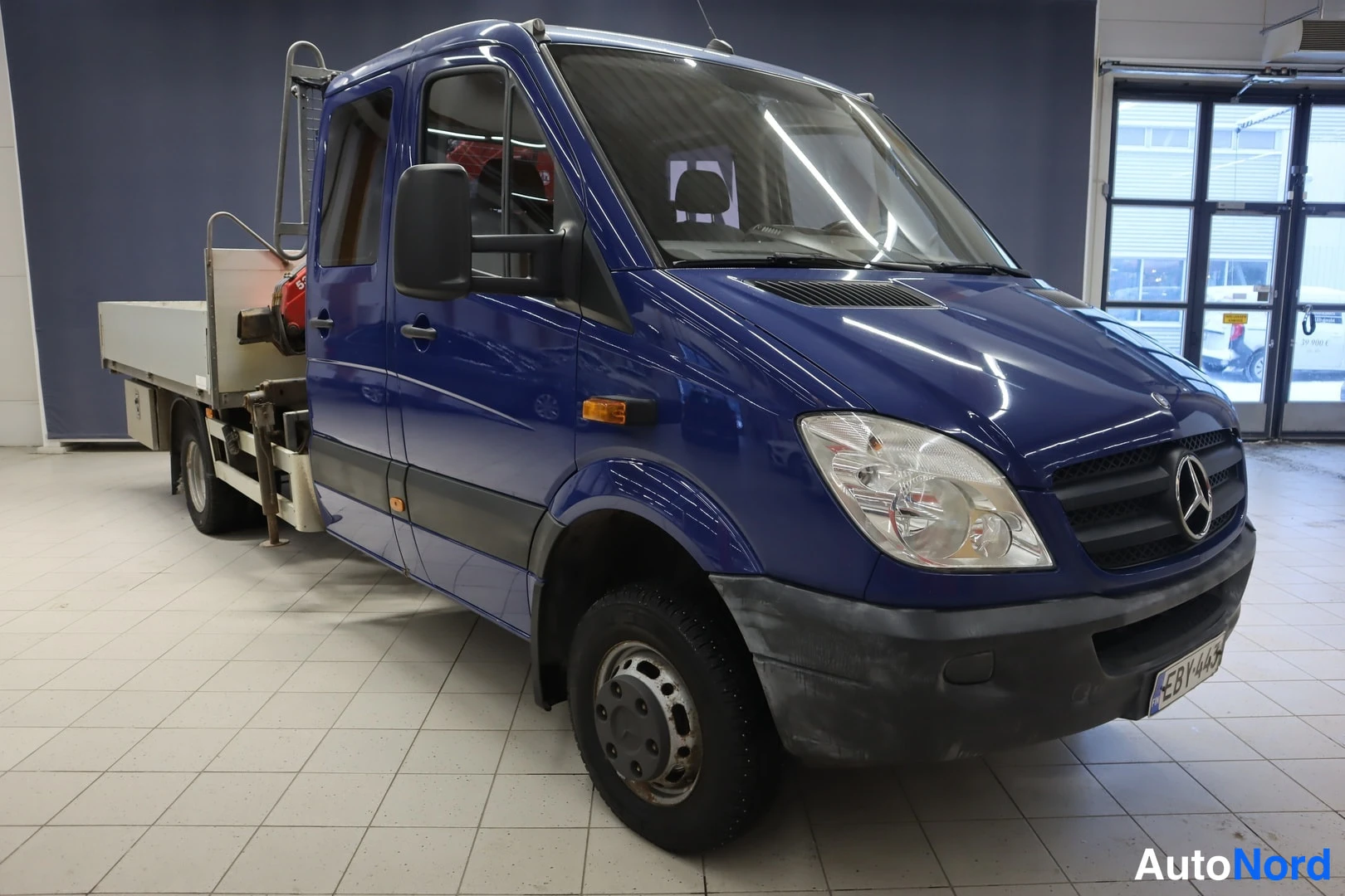 Etuviisto, nokka oikealle: Front three-quarter of a blue Mercedes-Benz truck in an indoor showroom.