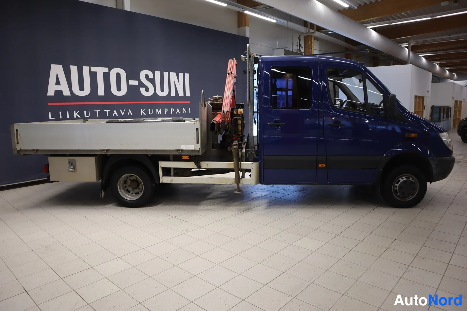 Sivulta: Side view of the blue truck with flatbed and crane inside a dealership hall with 'AUTO-SUNI' on the wall.