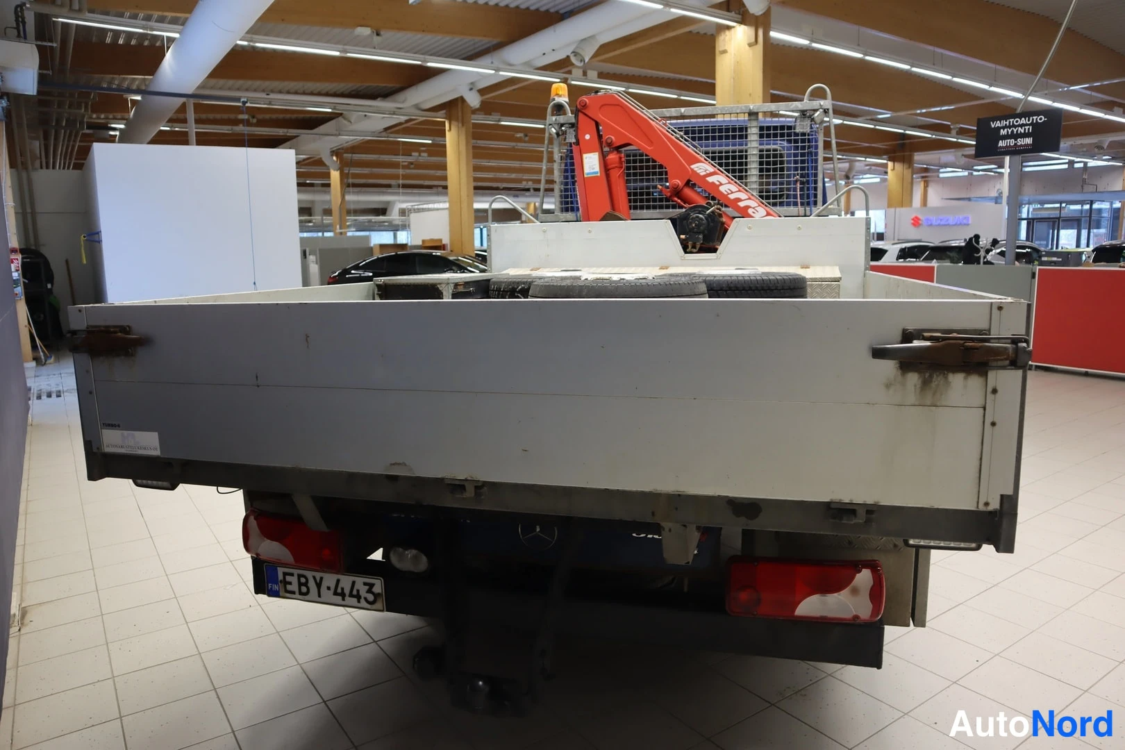 Takaa: Rear view of the white flatbed with a tire secured behind it and a spare wheel stored underneath the chassis. Red tail lights are visible on the bumper. Registr