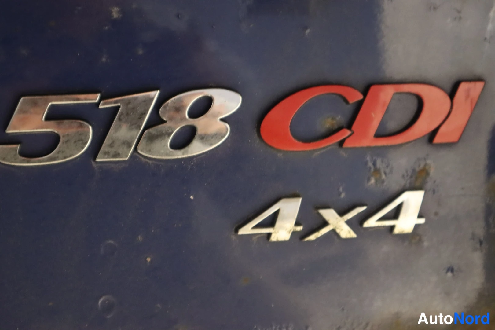 Muu: Lähikuva auton kyljen chromattu 518 CDI -merkki ja alla hopeiset 4x4-merkit tummansinisellä pinnalla.