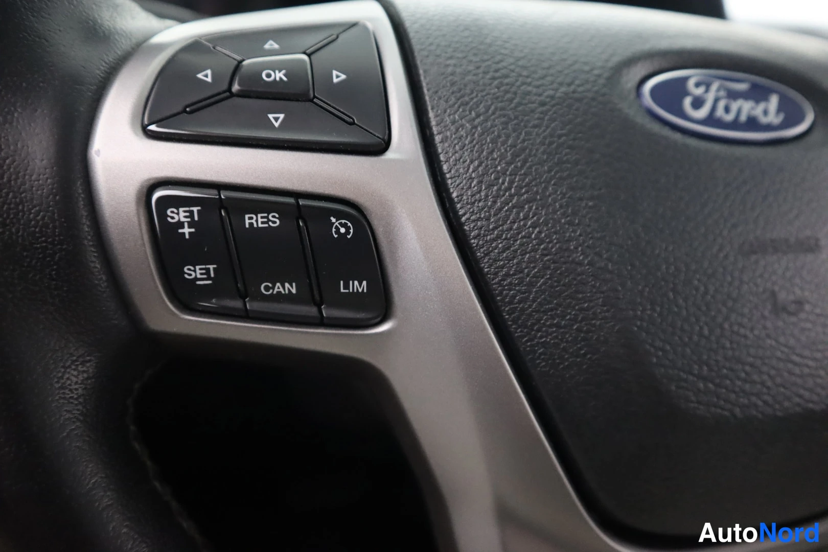 Sivulta: Ohjauspyörän vasemman puolen painikkeiden lähikuva: Ford-logo, OK-nuolipainikkeet sekä vakionopeudensäädön painikkeet SET+, SET-, RES, CAN ja LIM.