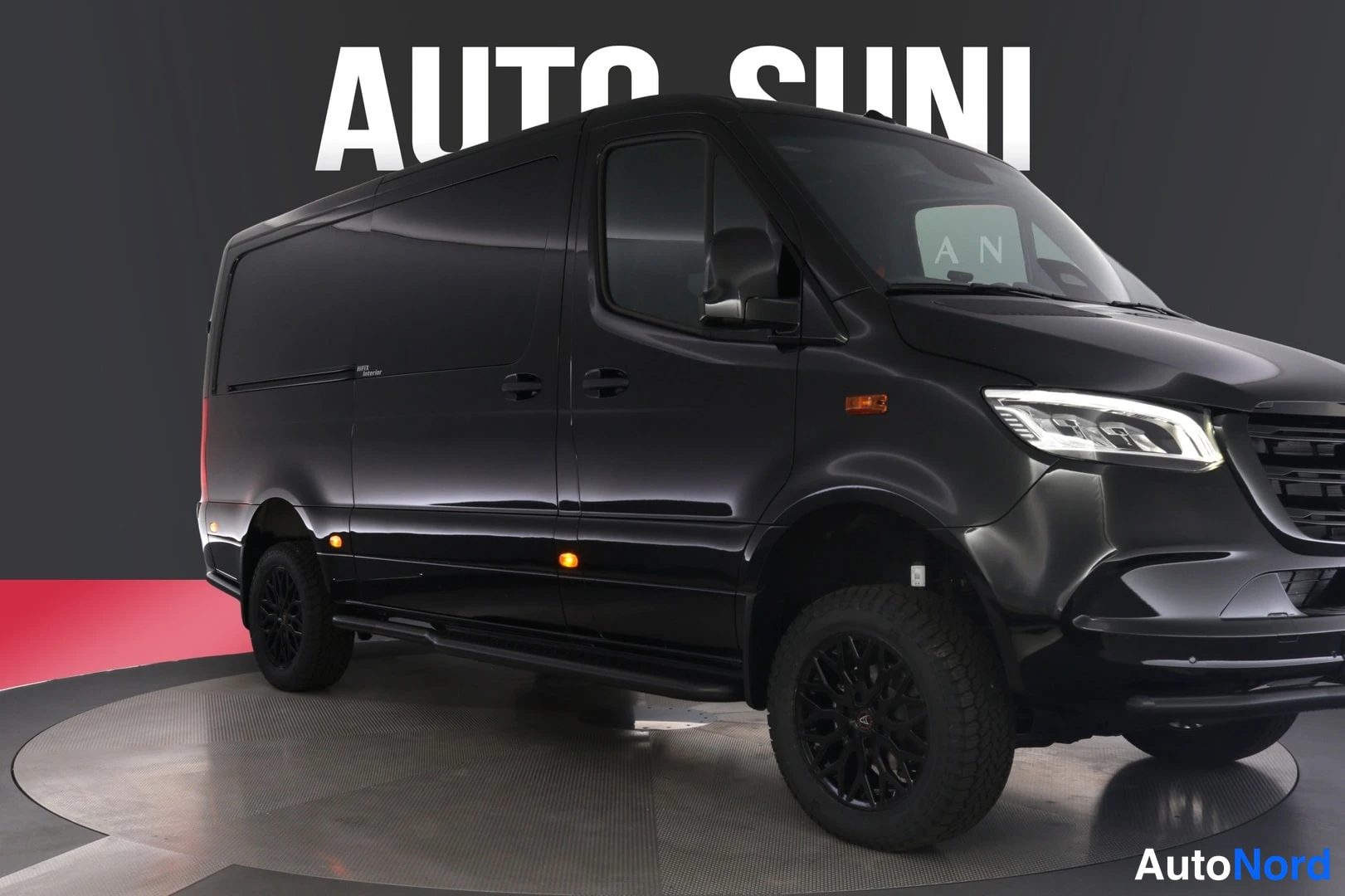 Etuviisto, nokka oikealle: Musta Mercedes-Benz Sprinter etuviistöstä oikealta kuvattuna showroomissa, tummat vanteet ja mustat kiinteät astinlaudat.
