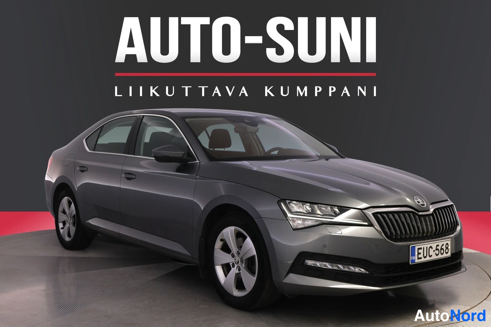 Etuviisto, nokka oikealle: Autoteen etukulmanäkymä: harmaa Škoda Superb -sedan, keulavälissä merkki ja rekisteritunnus, sivupeilit ja ovipaneelit näkyvissä.