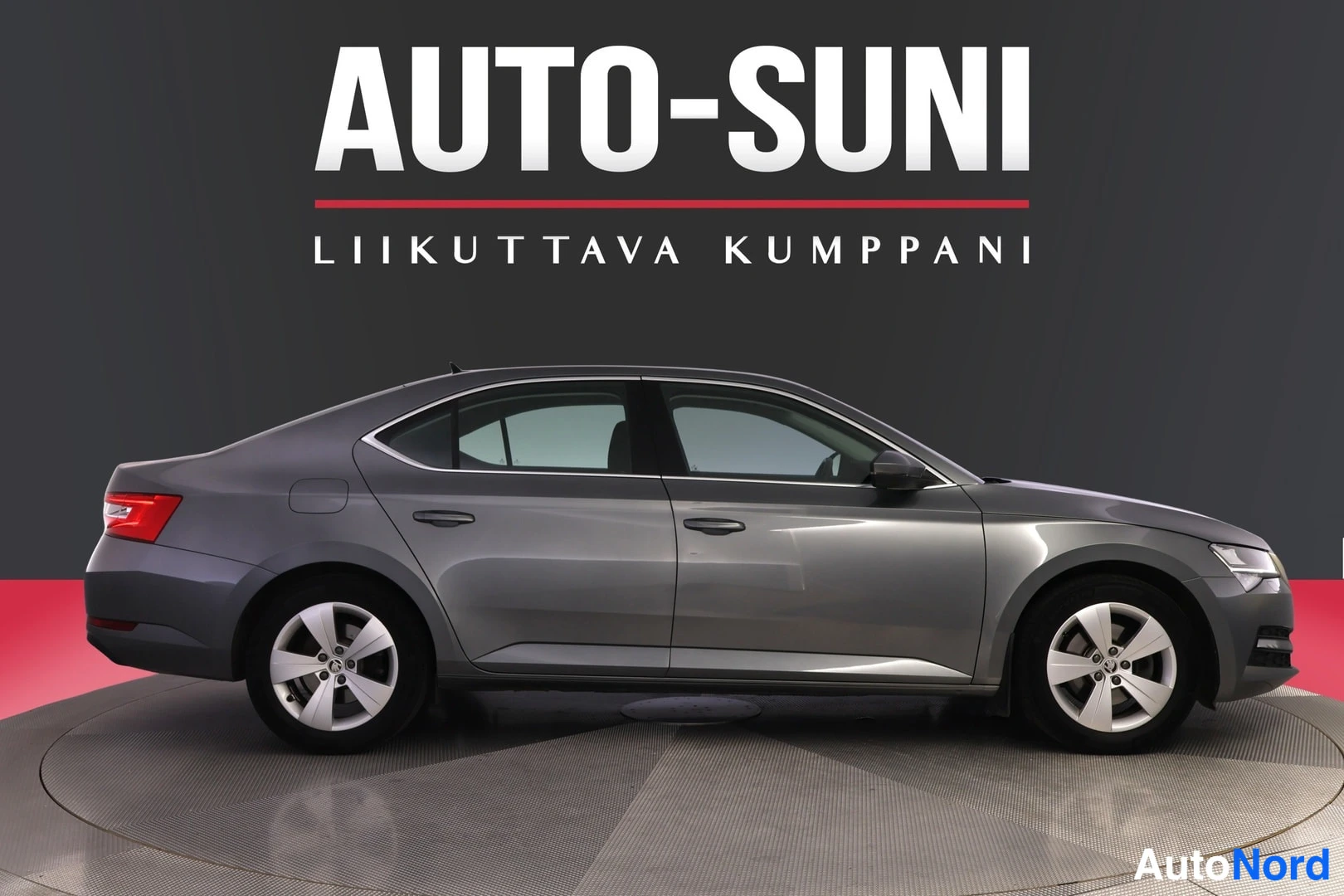 Sivulta: Autoteen sivunäkymä vasemmalta: harmaa Škoda Superb -sedan kattoo, ovista ja pyöristä valaistuna studioissa.