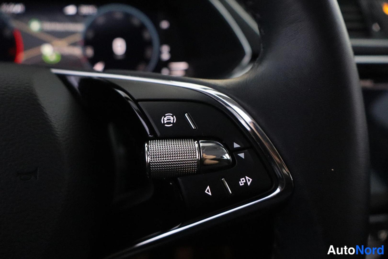 Ohjaamo: Close-up of the multifunction steering wheel controls on the left spoke. A silver scroll wheel is surrounded by buttons including a parking sensor icon.