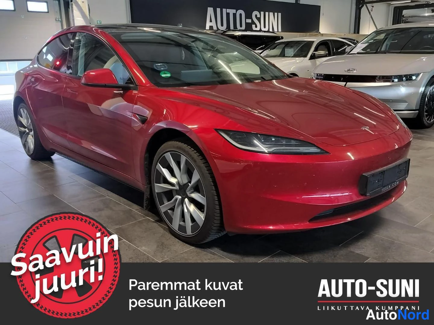 Etuviisto, nokka oikealle: Punainen Tesla Model 3 -henkilöauto etuosa vasemmalta kuvattuna autosalonin sisällä, muiden autojen taustalla.