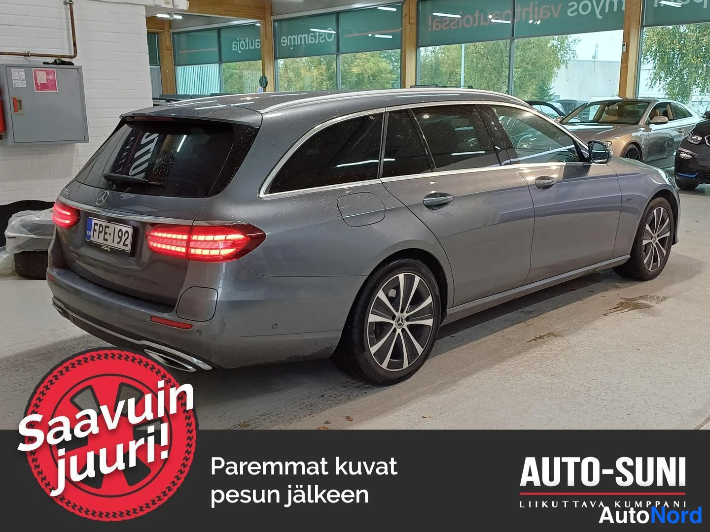 Takaviisto: Hopeanharmaa Mercedes-Benz E-farmari takaa sivulta hallissa, takavalot syttyneinä, rekisterikilpi.