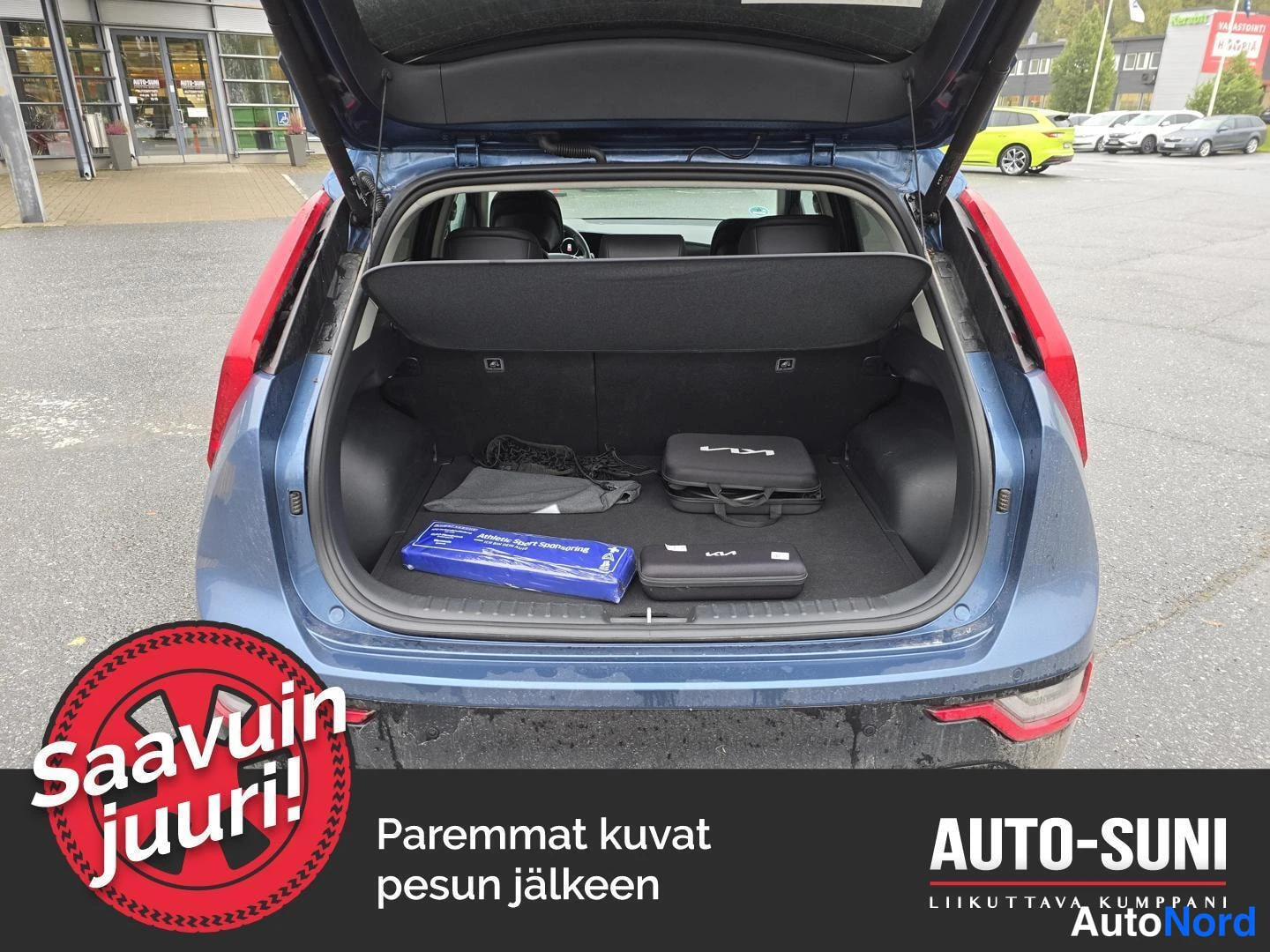 Takaa: Sinertävän harmaan auton avattu tavaratila takaapäin; tilassa lattiaverhot, laturi ja pussit.
