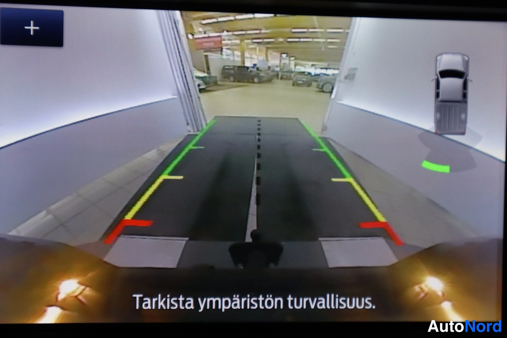 Yksityiskohta: Infotainment-näyttö näyttää peruutuskameran kuvan ohjausviivoineen ja ajoneuvon piirroksen; teksti 'Tarkista ympäristön turvallisuus.'