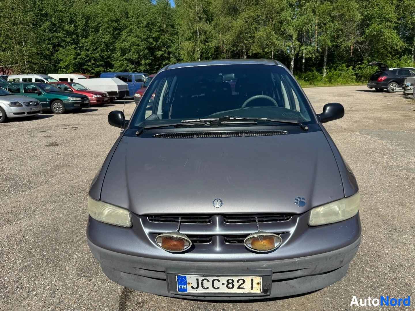 Edestä: Chrysler Voyagerin keula suoraan edestä kuvattuna: harmaa kori, tumma ohjaamo tuulilasin läpi, kaksi pyöreää lisävaloa etusäleikössä ja rekisterikilpi. Taustall