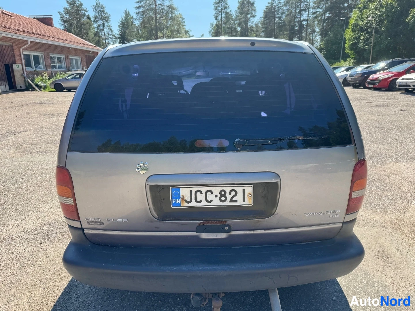 Takaa: Chrysler Voyagerin takaosa: rekisterikilpi, takaluukussa CHRYSLER- ja VOYAGER SE -tekstit; osa korista peittyy pölyyn. Kuva epätarkka.