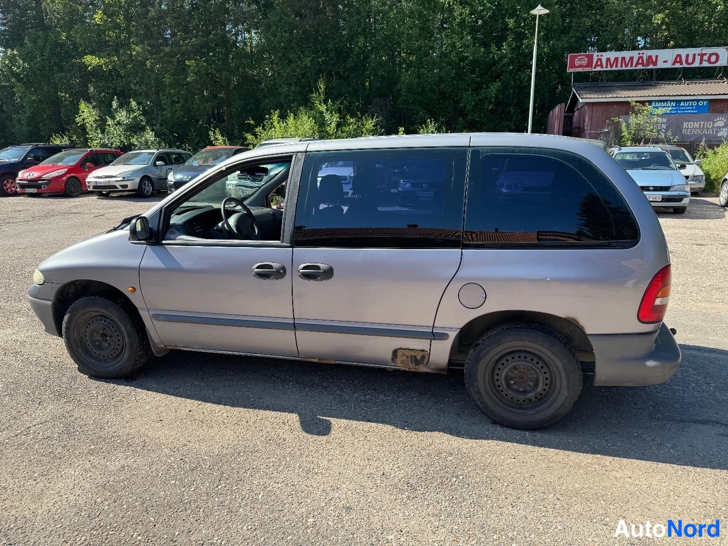 Sivulta: Side view of a silver Chrysler minivan parked outdoors on a gravel lot. The vehicle features dark tinted windows and black steel wheels without wheel covers. Ru