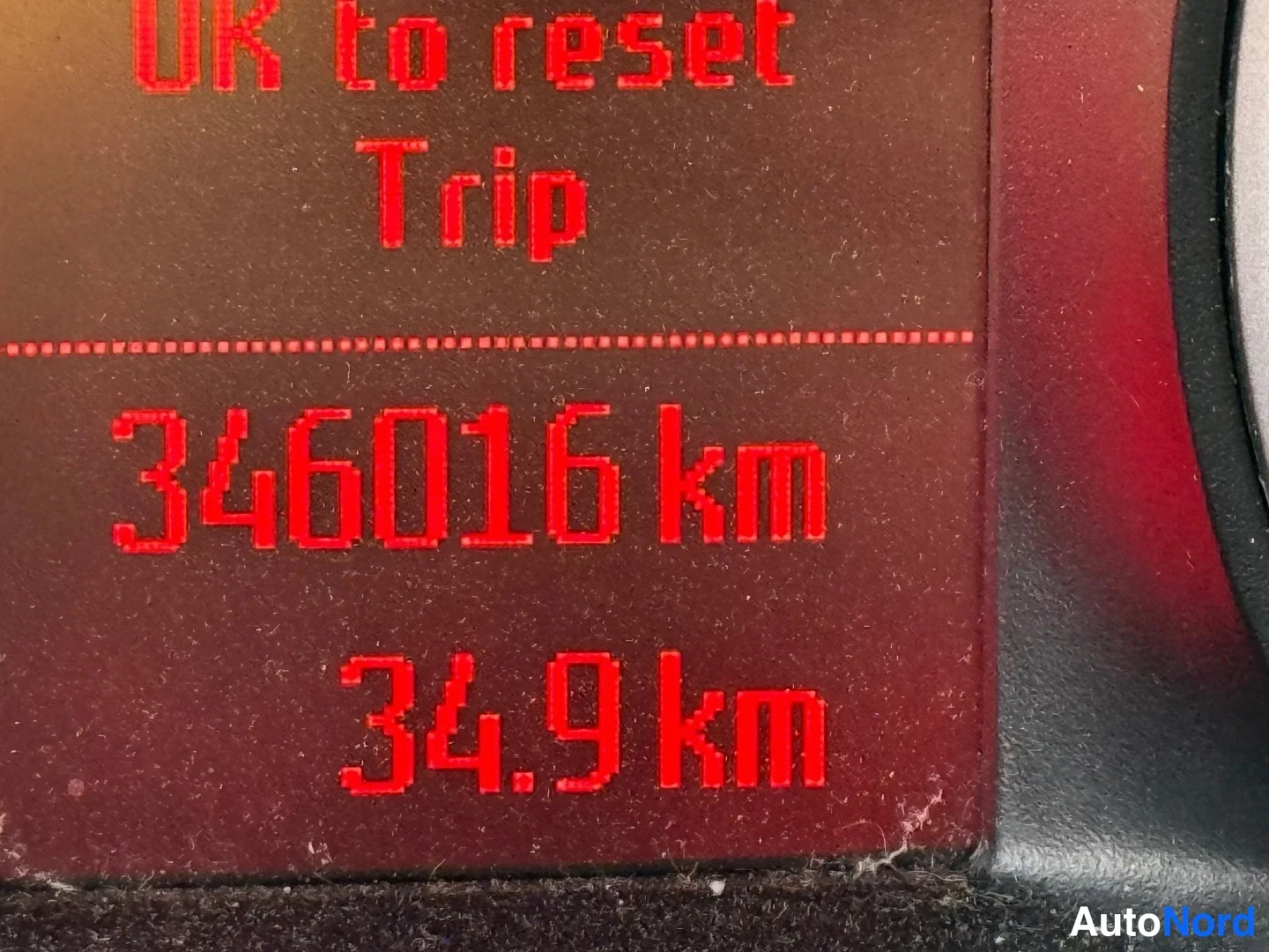 Yksityiskohta: Autojen mittaristön näyttö lähikuvassa, punainen teksti 'OK to reset Trip' ja lukema 346016 km sekä matkamittari 34,9 km.