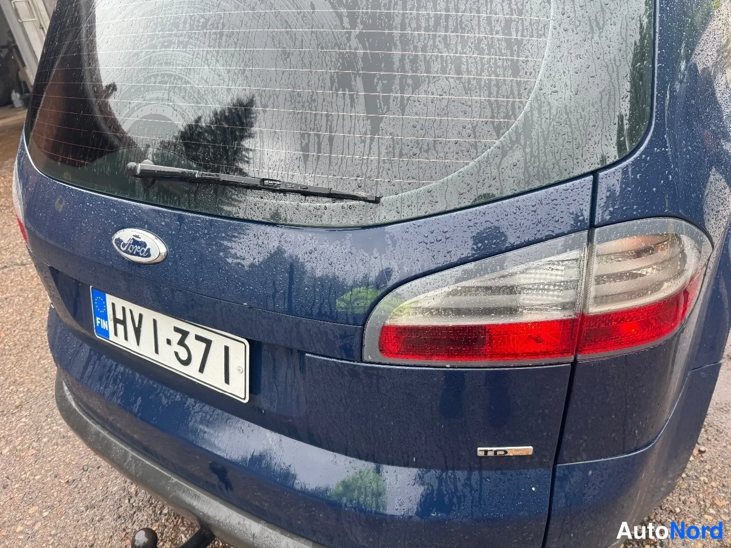 Takaa: Tummansinisen tila-auton takapää lähikuvassa: Ford-merkki, rekisterikilpi 'HV I-371' ja takavalot, pinta märkä sadevedestä.
