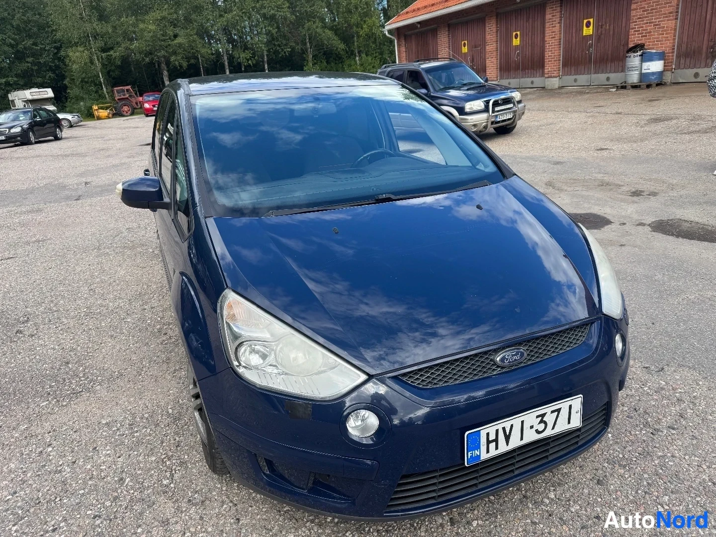 Etuviisto, nokka vasemmalle: Sininen Ford S-MAX kuvattu kolmannelta sektorilta edestä vasemmalta, etupaneeli ja ajokkinaapuri puoli näkyvissä hiekkaparkkipaikalla.