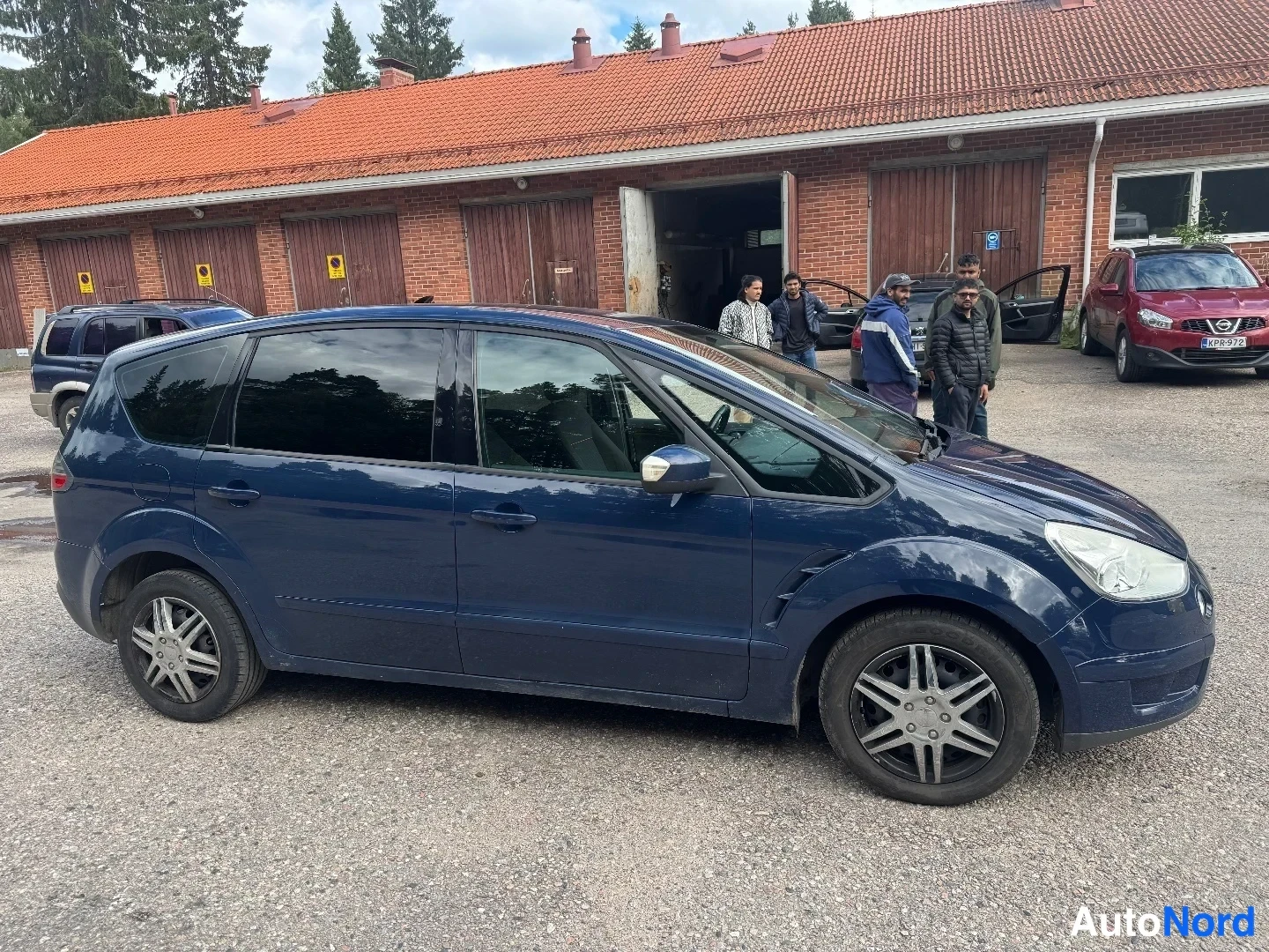 Etuviisto, nokka oikealle: Tummansininen tila-auto etuviistosta oikealta; kylki ovineen ja etupää ajovaloineen näkyvät.