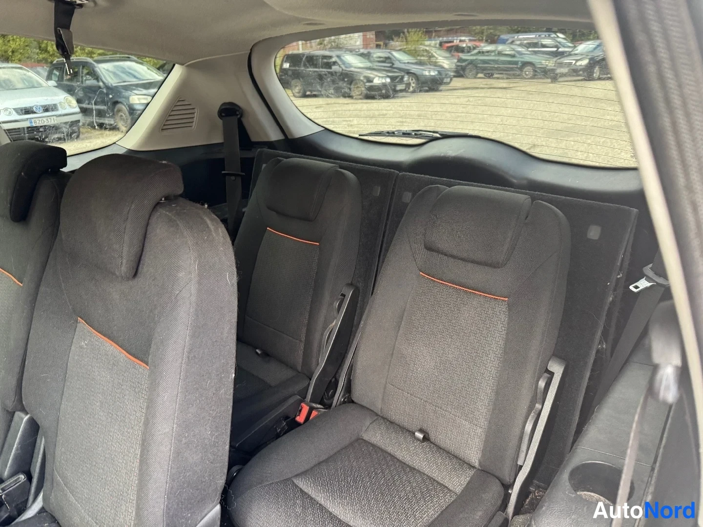 Muu: Auto sisältä kuvattuna: kolme erillistä takaistuinta, joiden verhoilu on tummaa kangasta oransseilla ompeleilla; taustalla takaluukun sisäpinta, lasi ja pyyhin.