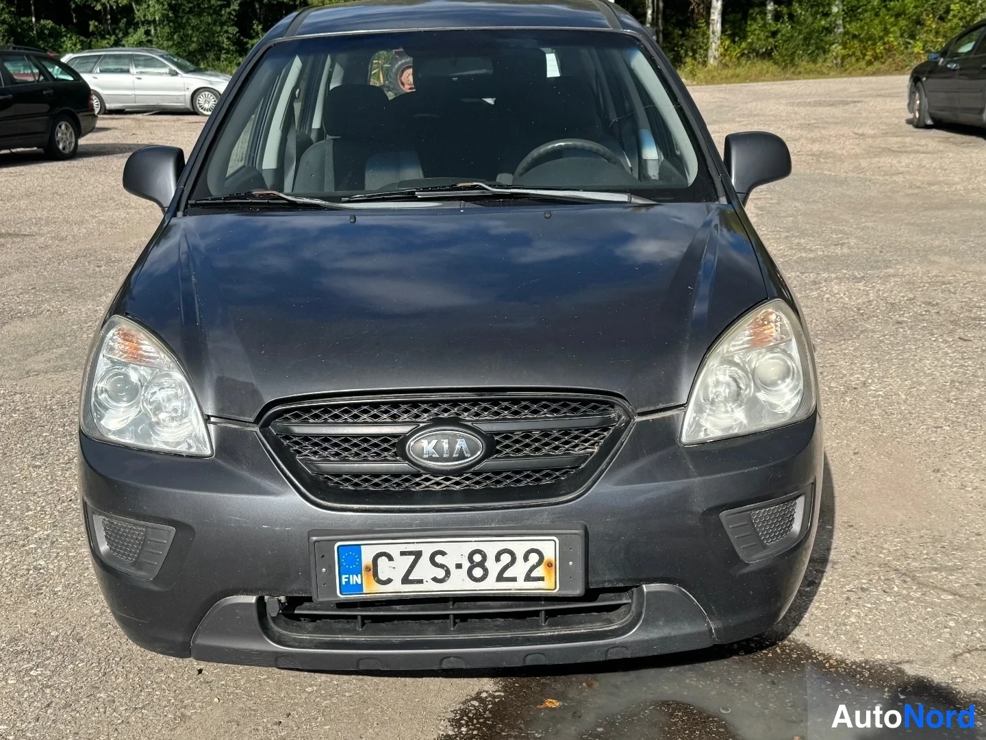 Edestä: Tummanharmaa Kia Carens kuvattuna suoraan edestä: KIA-merkki mustassa etusäleikössä, sumuvaloaukot etupuskurissa ja rekisterikilpi.