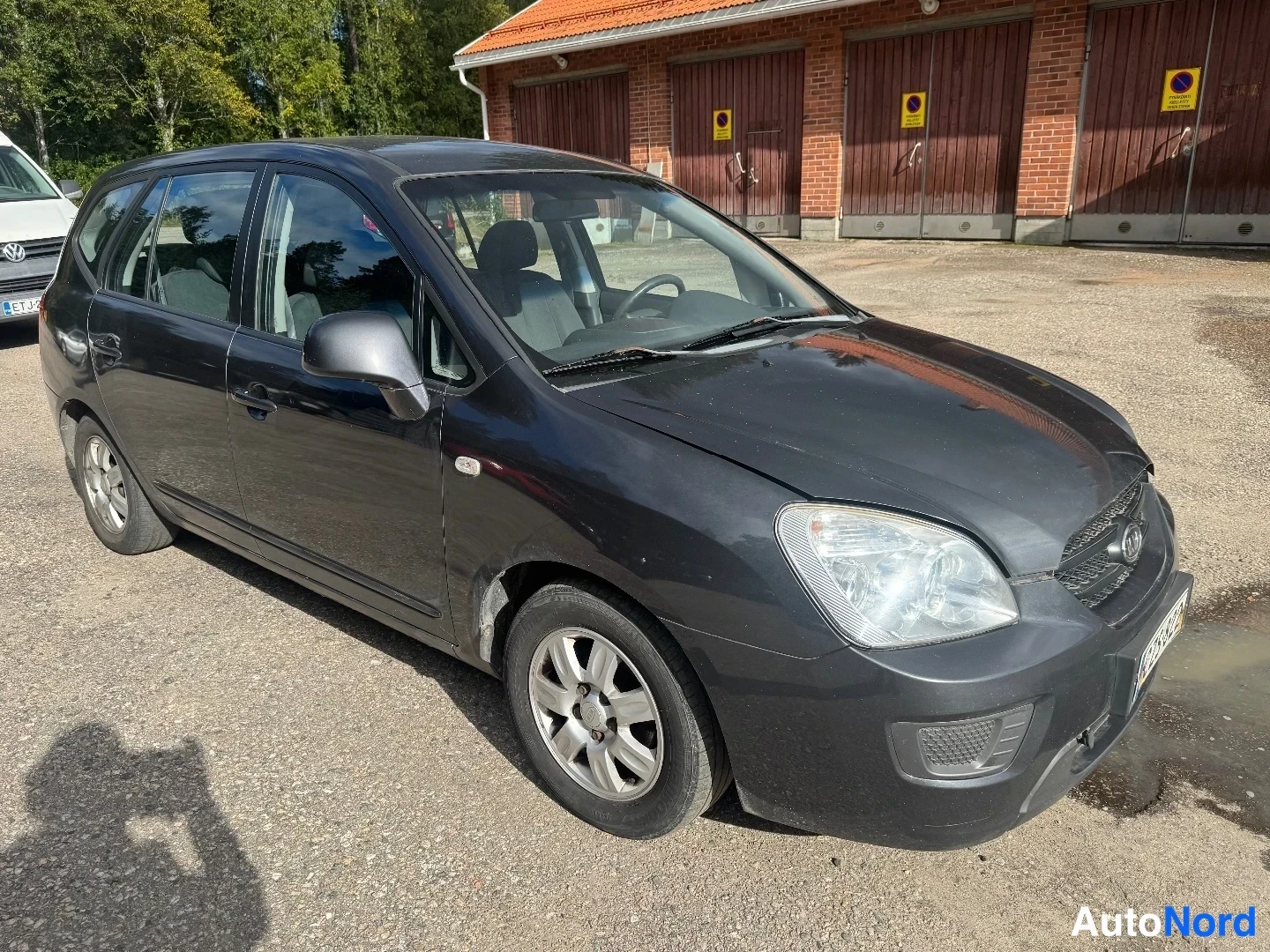Etuviisto, nokka oikealle: Auton etukolmasosa kuvattuna keula oikealla; tummanharmaa farmarikorinen henkilöauto seisoo sorapihalla tiilirakennuksen edessä.