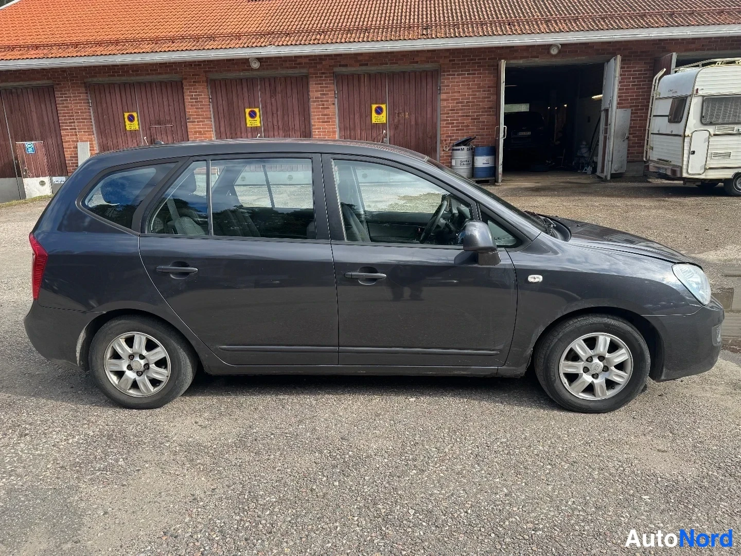 Etuviisto, nokka oikealle: Tummanharmaa farmarityyppinen henkilöauto hieman viistosti sivulta kuvattuna, keula oikealla; taustalla punatiilinen varastorakennus.