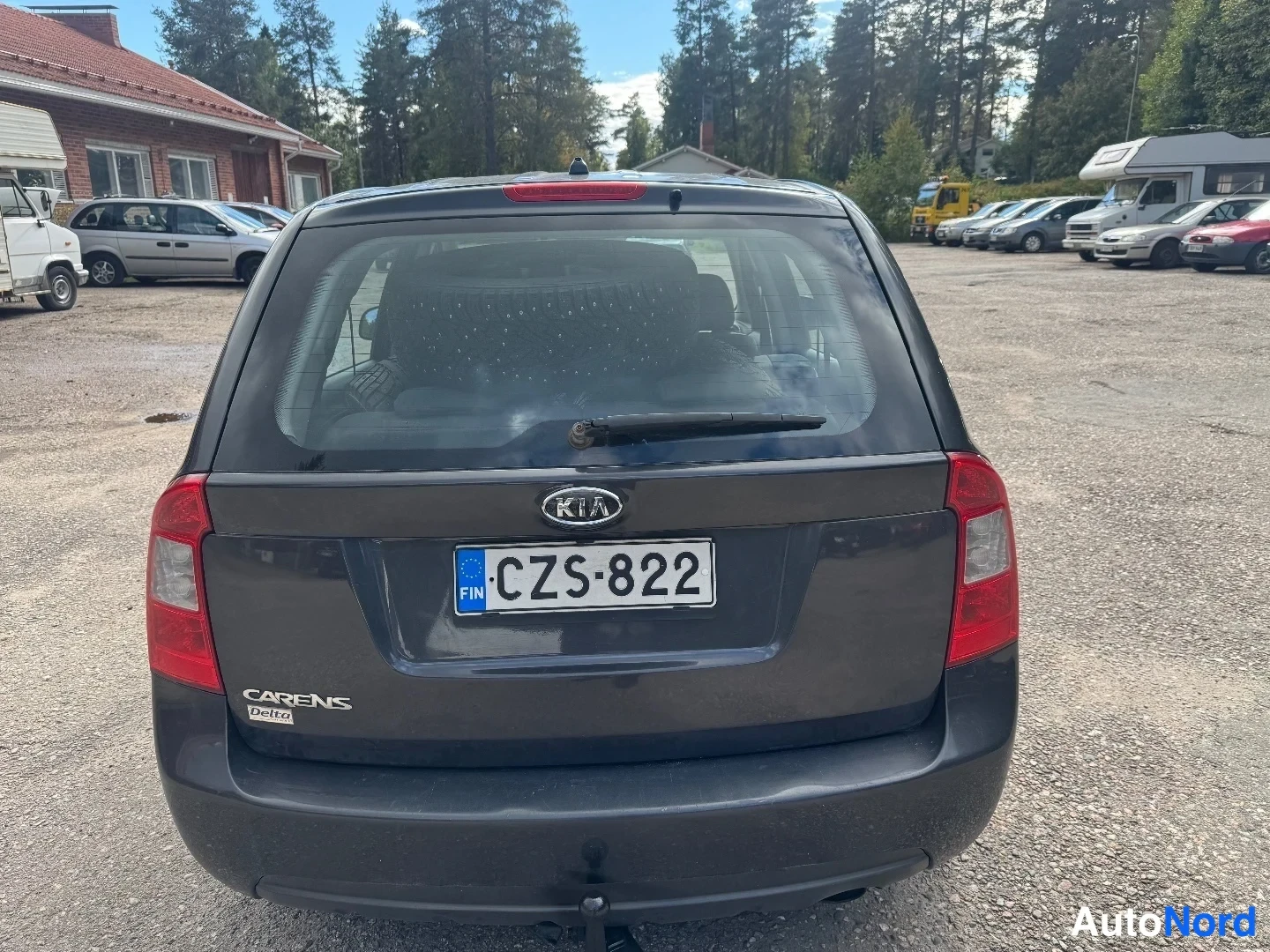 Takaa: Kia Carensin takaosa suoraan takaa kuvattuna: KIA-logo ja CARENS Delta -teksti takaluukussa, rekisterikilpi, punaiset takavalot molemmin puolin ja ajopyyhkijä t