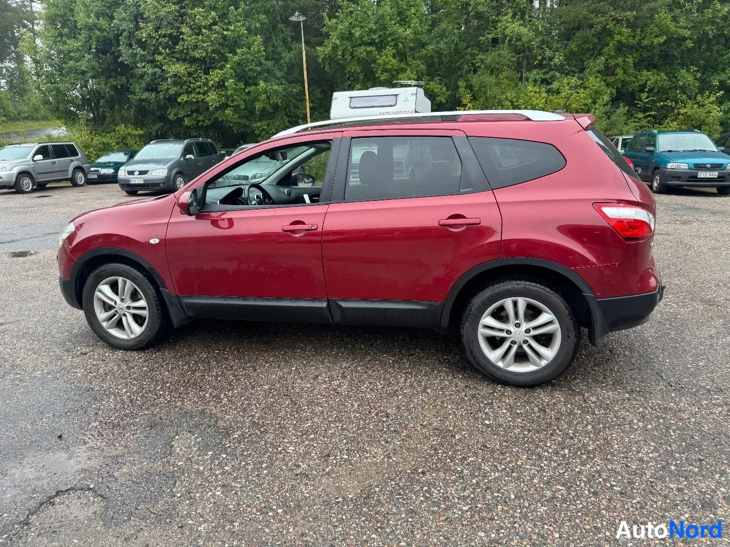 Takaviisto: Punainen farmuritu SUV sivulta-takaa kuvattuna soraparkkipaikalla, takavalot ja hopeiset vanteet näkyvissä.
