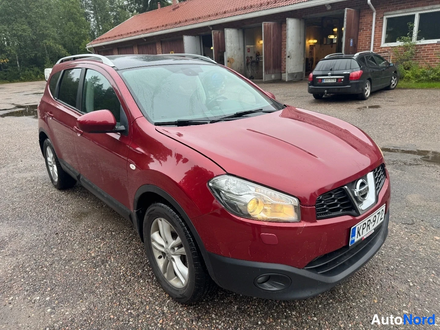 Etuviisto, nokka oikealle: Tummanpunainen henkilöauto etuviistosta, keula kuvan oikeassa laidassa. Rekisterikilpi, kattokaiteet ja kevytmetallivanteet näkyvät. Taustalla tiilinen a