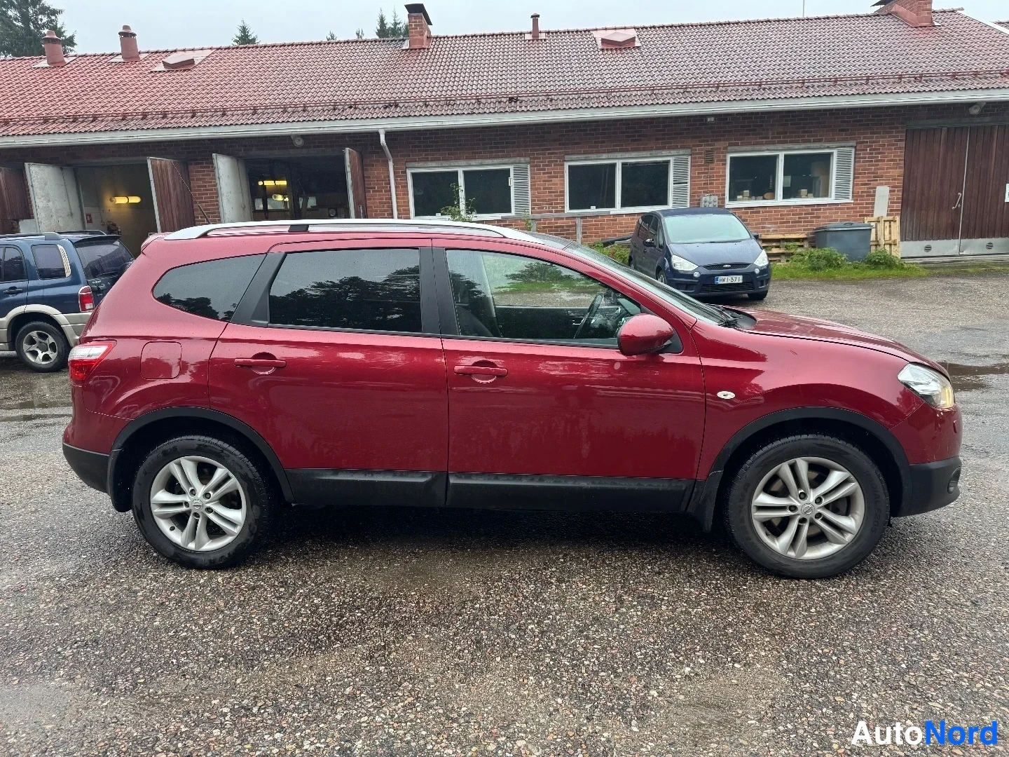 Sivulta: Punainen Nissan Qashqai -maastomalli seisoo asfaltilla. Autosta näkyy oikea kylki ja takana punatiilinen rakennus sekä toinen auto.