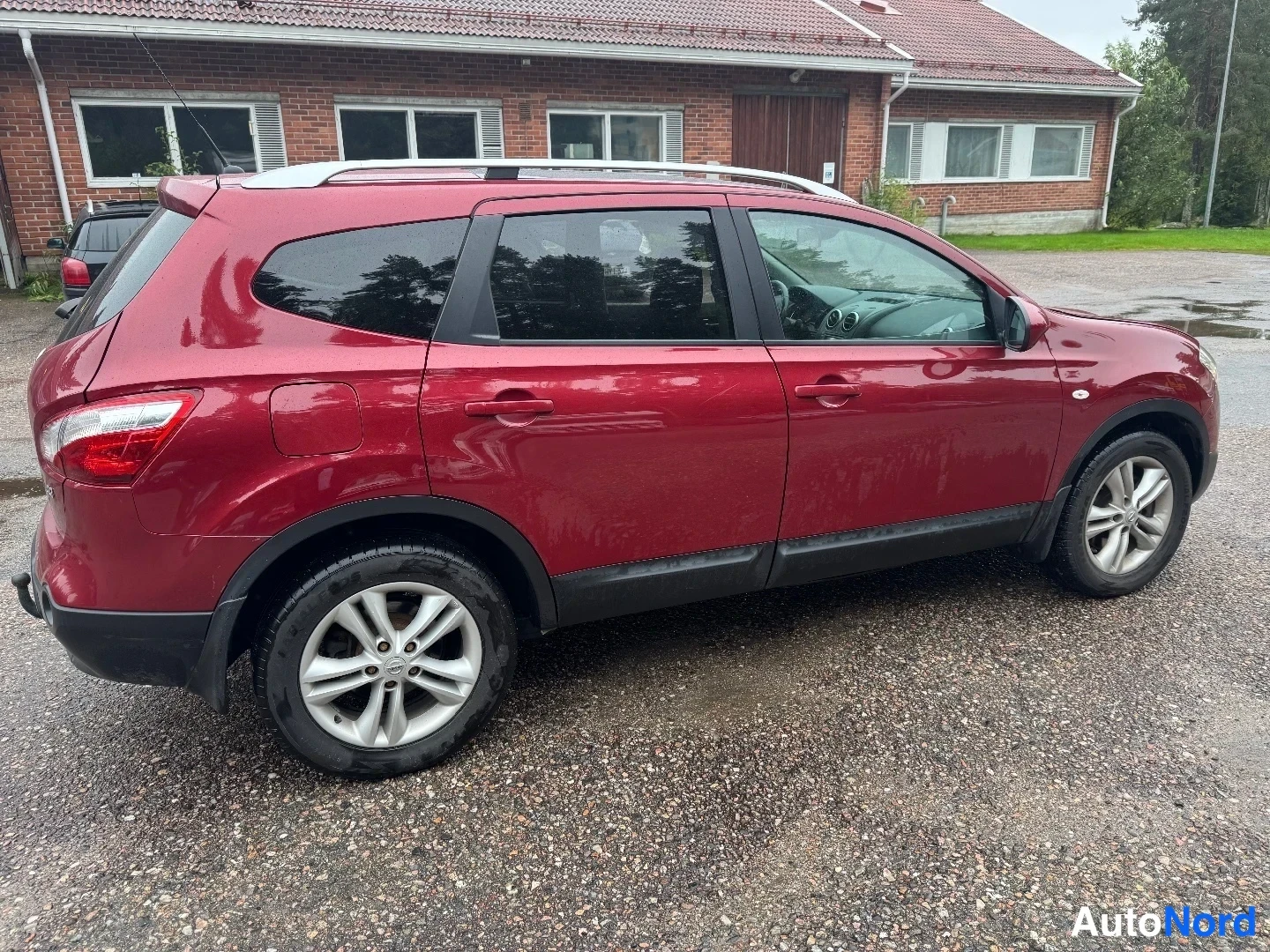 Takaviisto: Tummanpunainen Nissan Qashqai+2 kuvattuna takaviistosta vasemmalta: takaosa ja vasen kylki näkyvissä, kattokaiteet ja kevytmetallivanteet; taustalla tiilinen ra